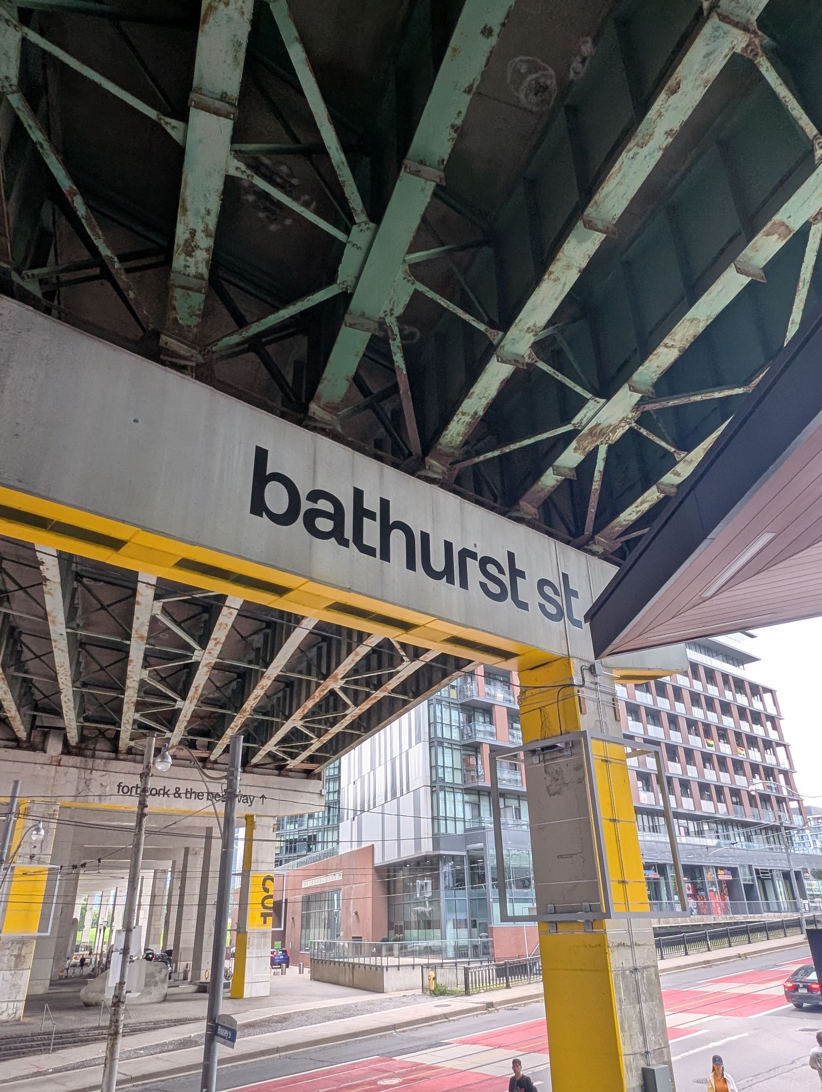 Looking up at the underside of the Gardiner Expressway overpass at Bathurst Street, with a black lowercase &ldquo;bathurst st&rdquo; sign in TBW Galeria mounted on a concrete beam, next to columns painted Bentway yellow.